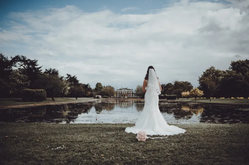 A bride in a lace fit and flare gown stands with her back to the camera by a tranquil lakeside with a grand manor house in the distance.