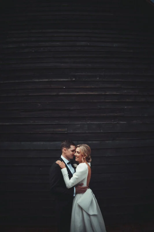 A bride and groom embracing in front of a black barn.