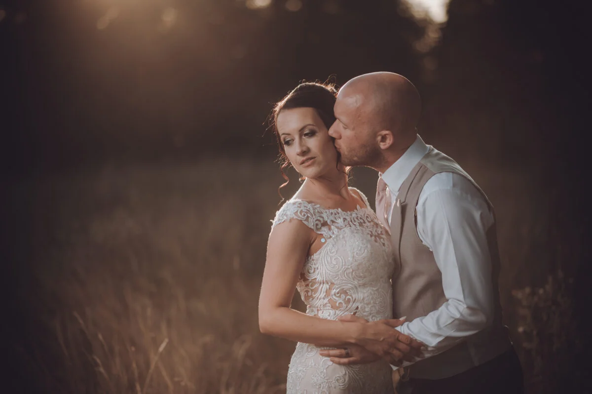 A groom gently kisses the bride's cheek outdoors during golden hour while she wears an intricate lace wedding dress with cap sleeves.