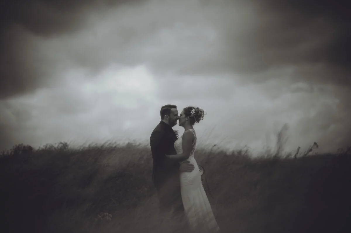A bride and groom standing in a field under a stormy sky.