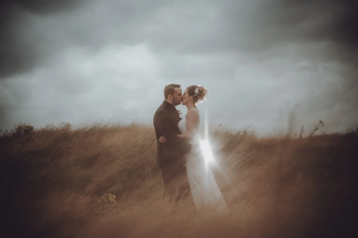 A bride and groom kissing in a field under a stormy sky.