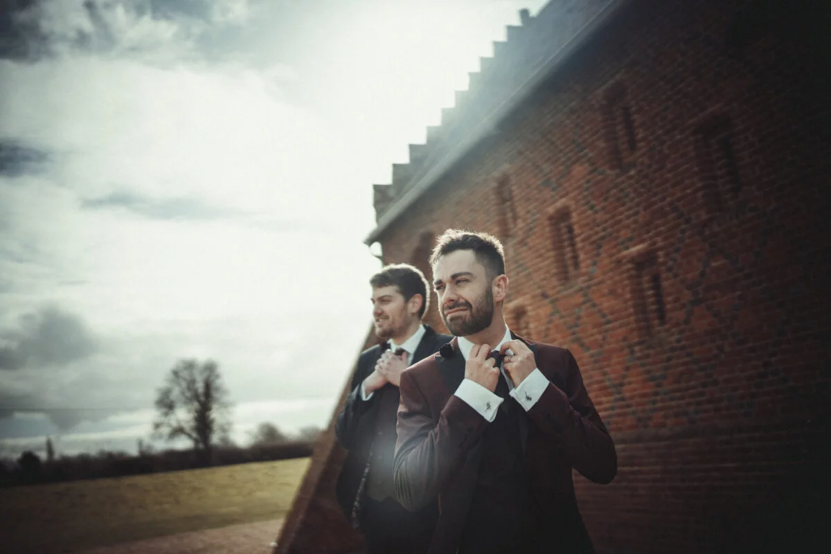 Two men tying their ties in front of a brick wall.