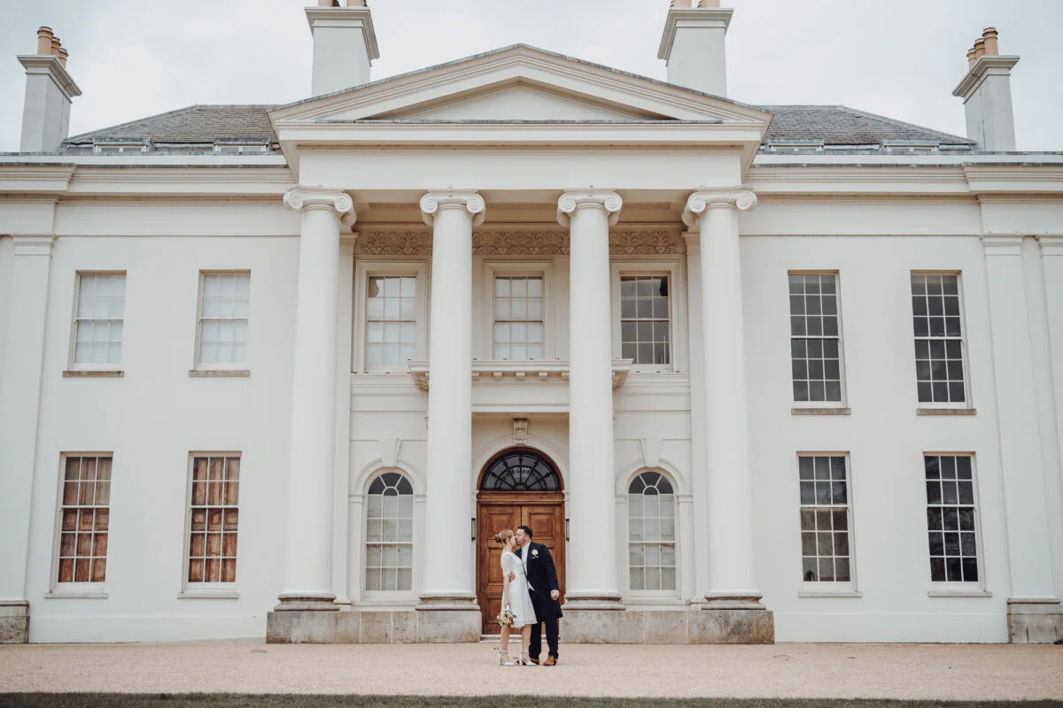 A bride and groom standing in front of a white mansion.