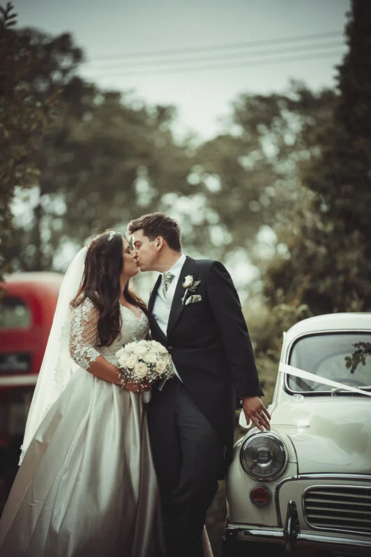 A bride and groom kiss in front of a classic car.