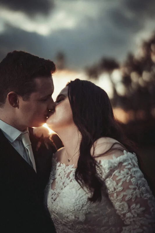 A bride and groom kissing in front of a sunset.