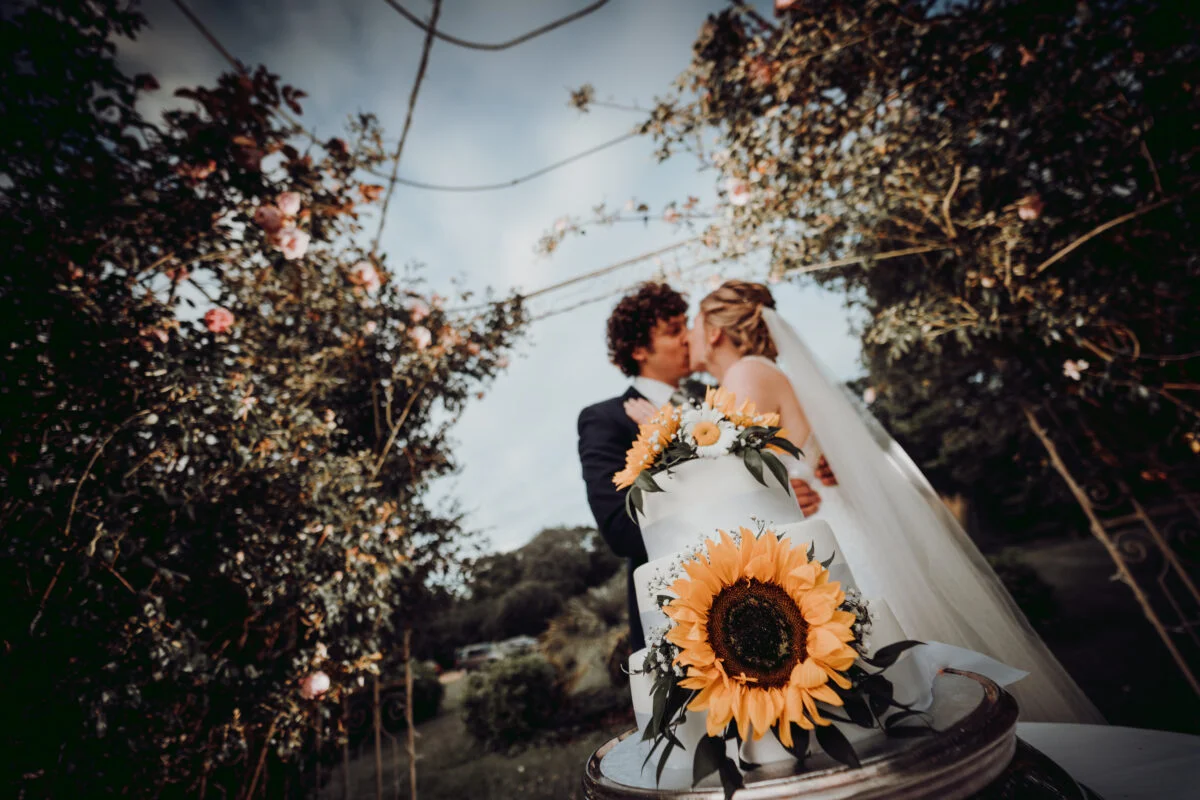 A bride and groom standing next to a wedding cake with sunflowers.
