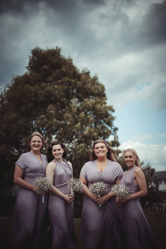 Bridesmaids in lavender dresses standing in front of a cloudy sky.