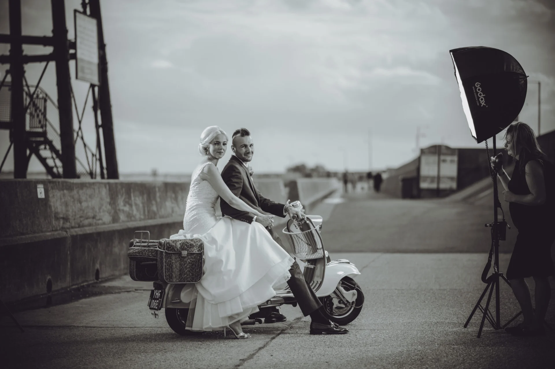 A bride and groom on a scooter in front of a bridge. (listing)