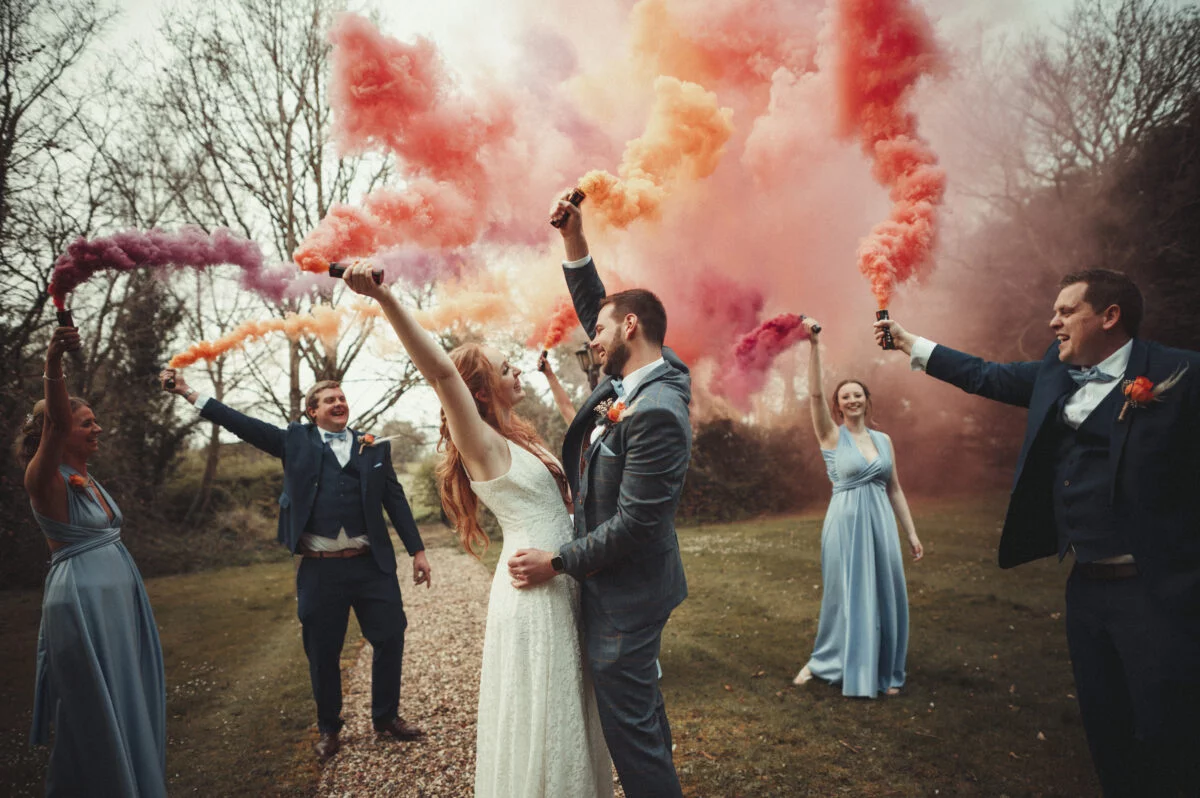A group of bridesmaids and grooms holding colored smoke bombs.