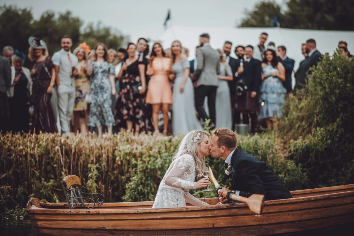 A bride and groom kissing in a boat in front of a crowd.