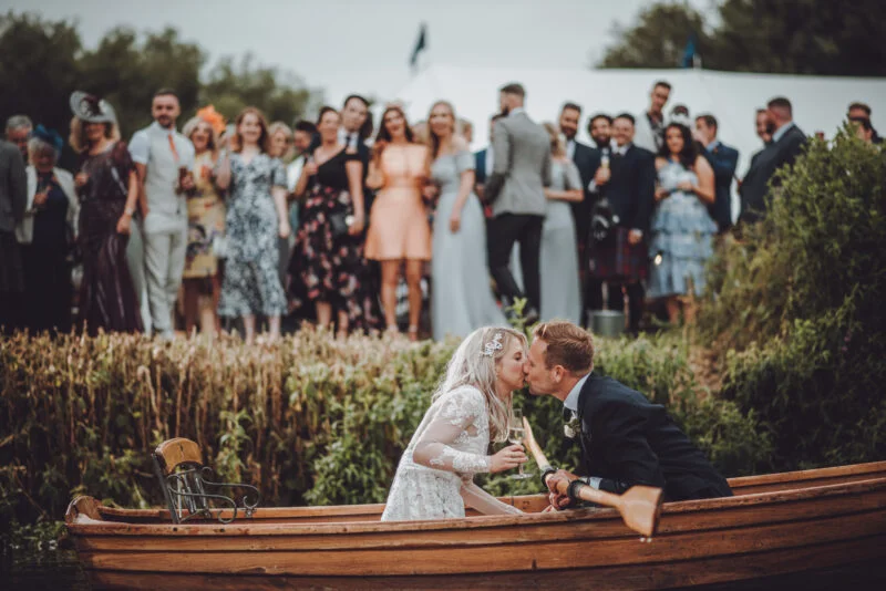 A bride and groom kissing in a boat in front of a crowd.