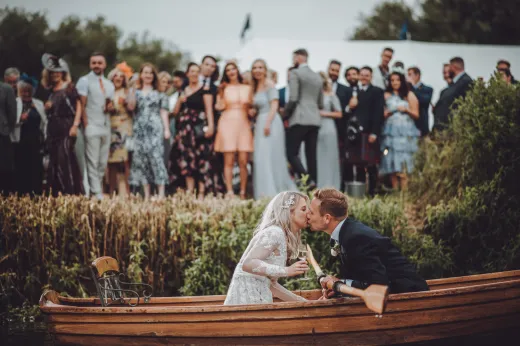 A bride and groom kissing in a boat in front of a crowd.