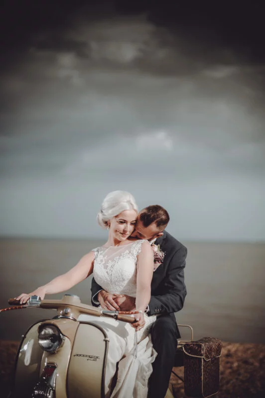 A bride and groom sitting on a vespa on the beach.