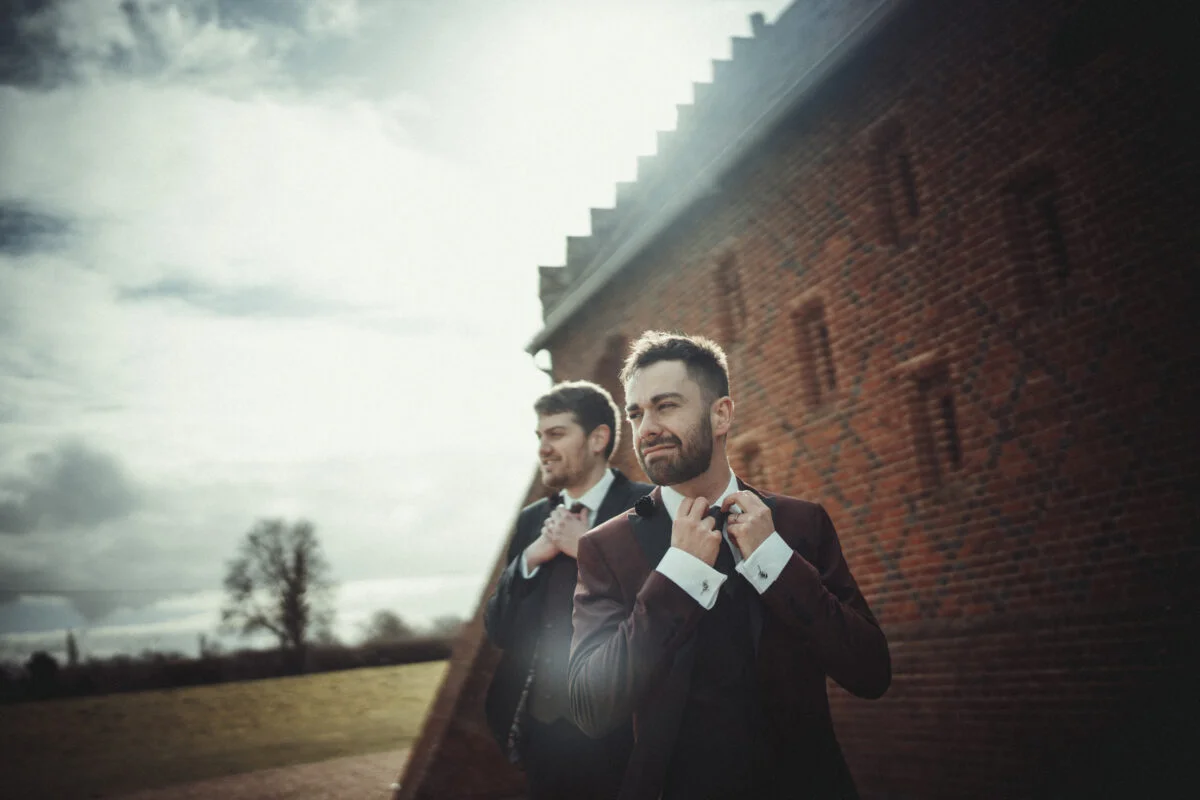 Two men tying their ties in front of a brick wall.