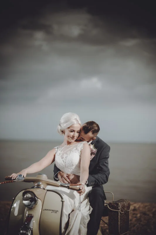 A bride and groom sitting on a vespa on the beach.