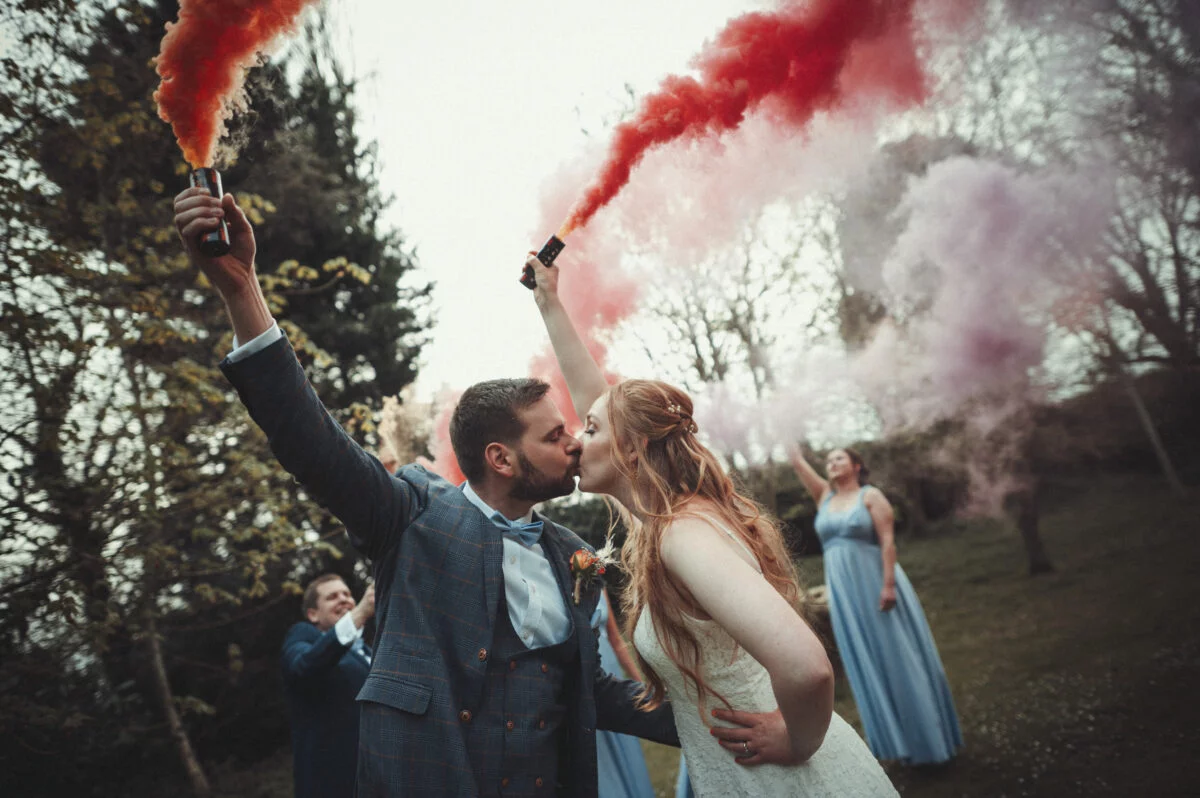 A bride and groom kissing with red and blue smoke bombs.