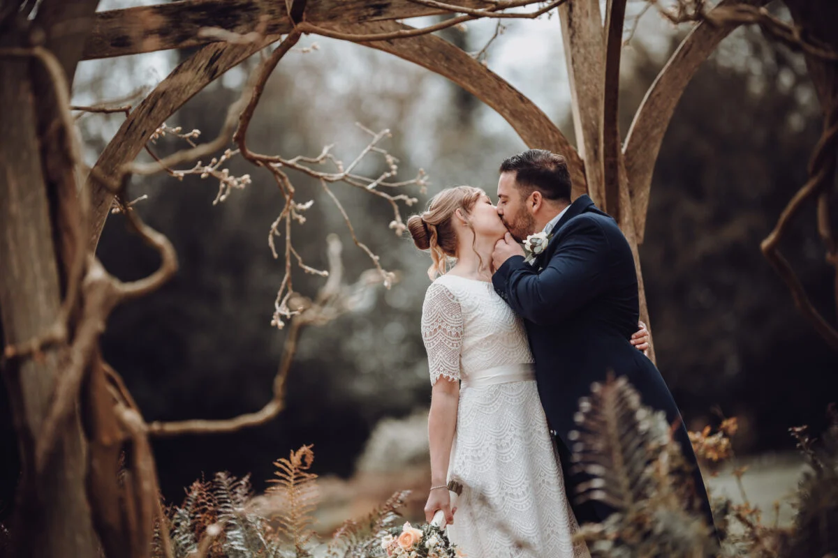 A bride and groom kissing under an arbour in the woods.