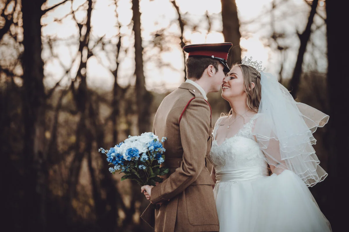 A newlywed couple shares a tender kiss in a woodland setting during golden hour, with the groom wearing a military uniform and holding a blue floral bouquet behind his back while the bride wears a tiara and veil.