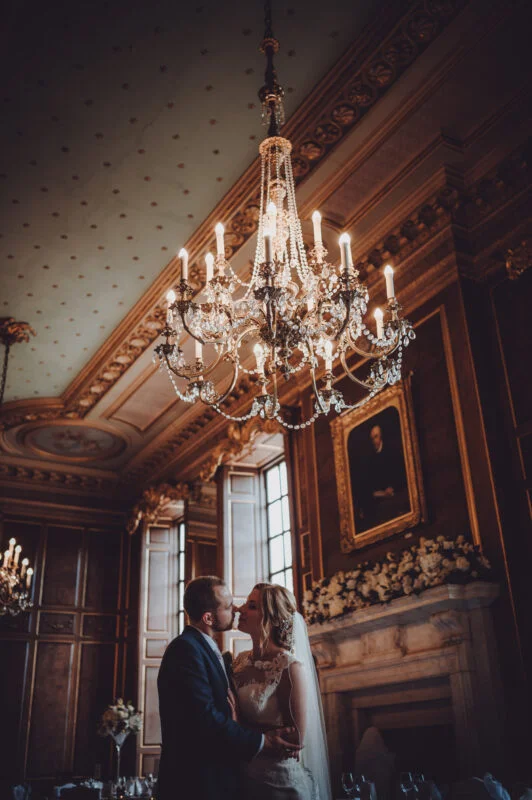A bride and groom kiss in front of a chandelier.