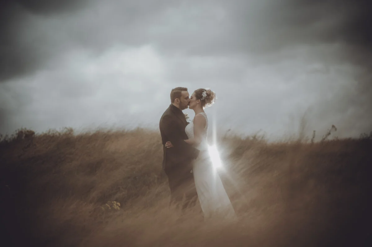 A bride and groom kissing in a field under a stormy sky.