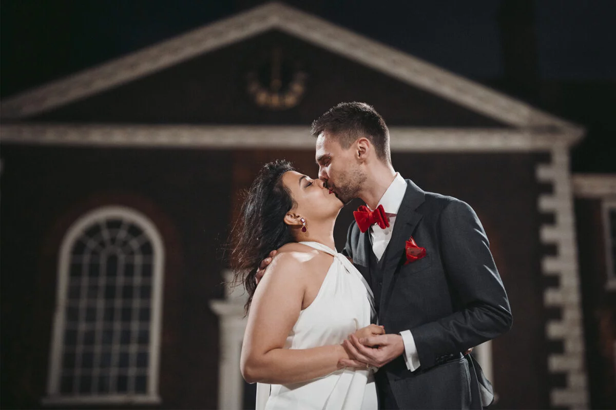 A bride and groom kiss in front of a large building at night.