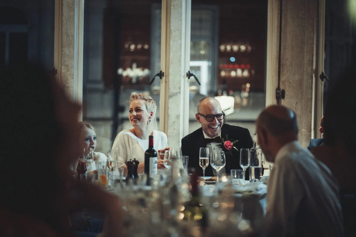 A bride and groom share a joyful moment laughing together at the top table during wedding speeches, viewed through a window.