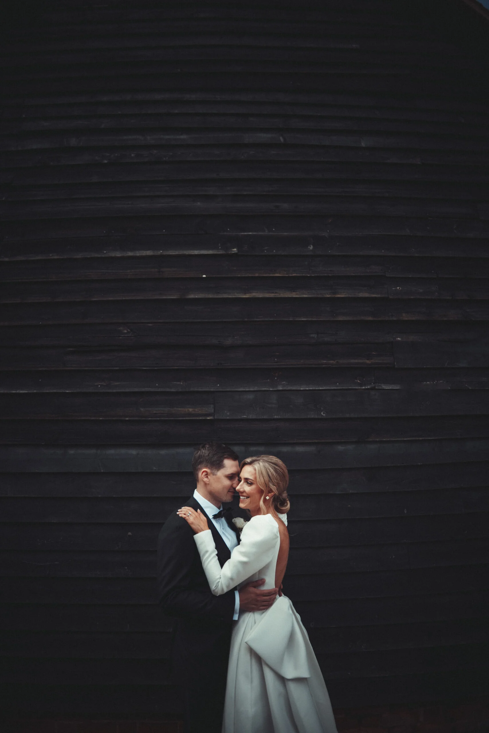 A bride and groom embracing in front of a black barn. (listing)