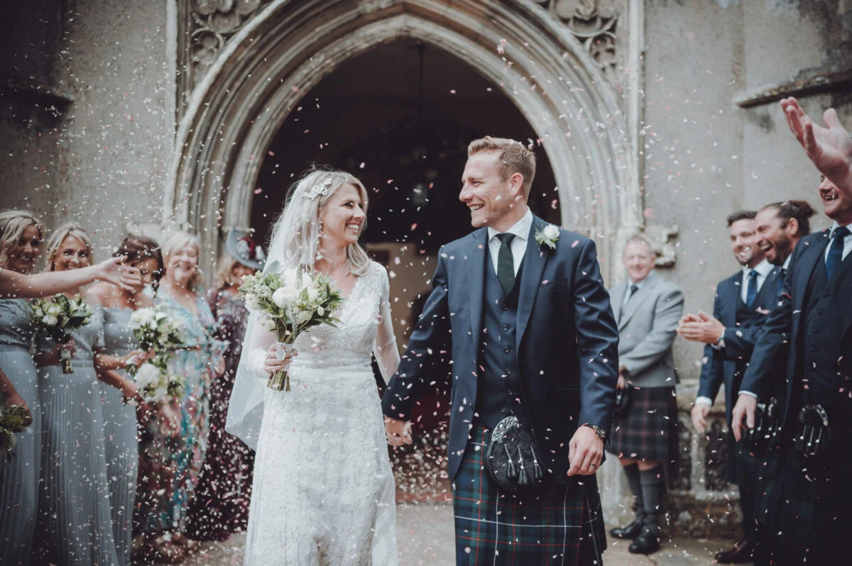 A bride and groom walk out of a church with confetti thrown at them.