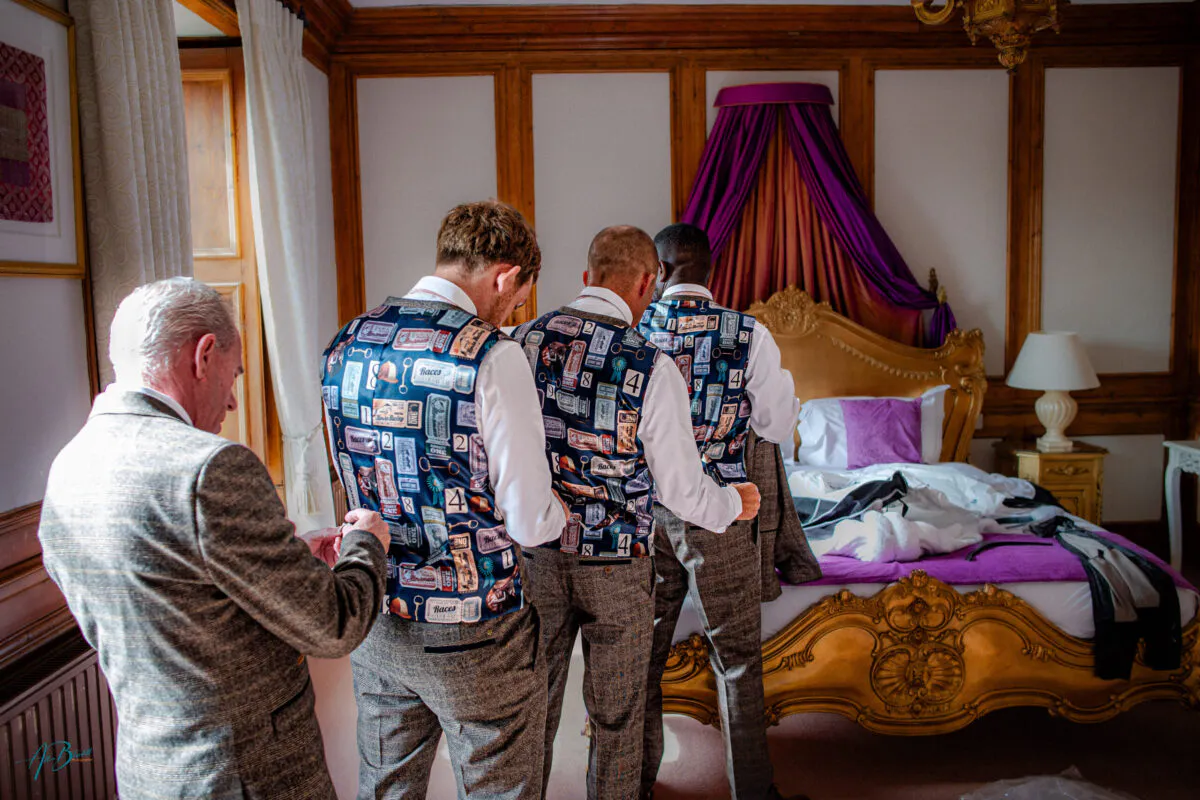 Three groomsmen and an older guest stand with their backs to the camera inside a wood-panelled bedroom, wearing distinctive patterned waistcoats as they finish getting ready for the wedding.