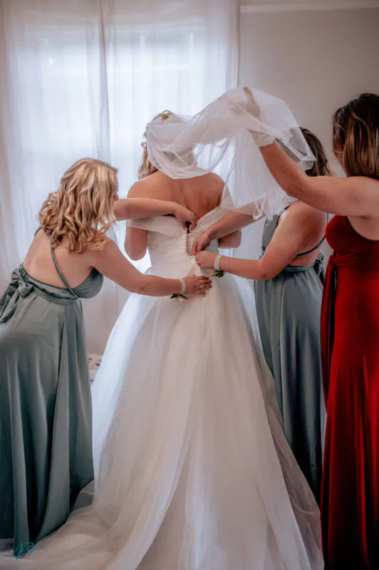 Three bridesmaids assist the bride with buttoning her white wedding dress and placing her veil during morning preparations.