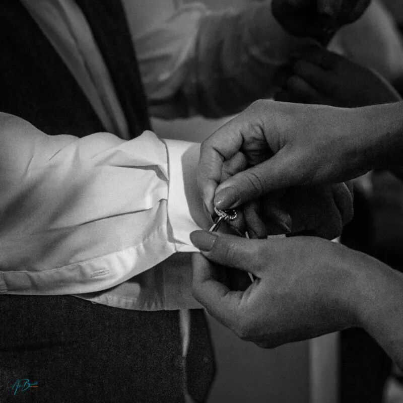 A close-up black and white photograph captures someone carefully fastening a cufflink onto the white shirt sleeve of a groom during morning preparations.