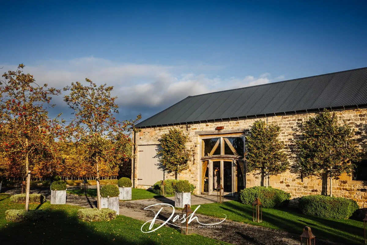 An exterior view of a rustic stone barn wedding venue bathed in golden autumn sunshine under a clear blue sky.