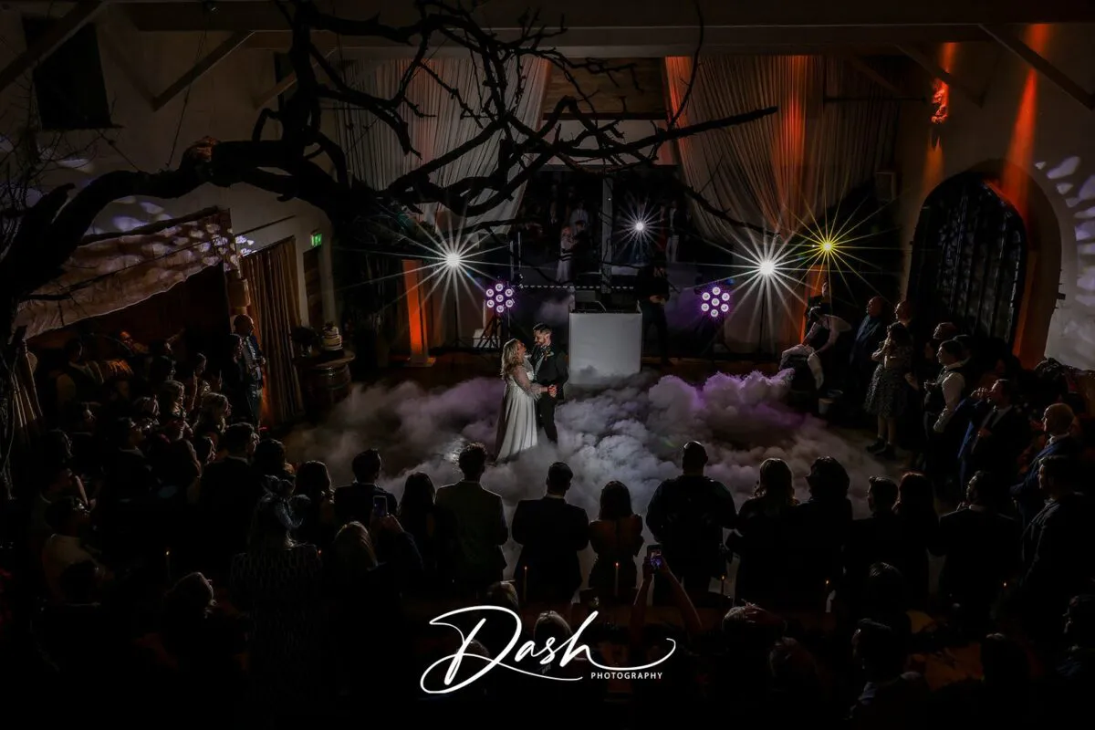 A newlywed couple shares their first dance on a dance floor filled with low-lying smoke inside a rustic barn illuminated by dramatic stage lights and surrounded by watching guests.
