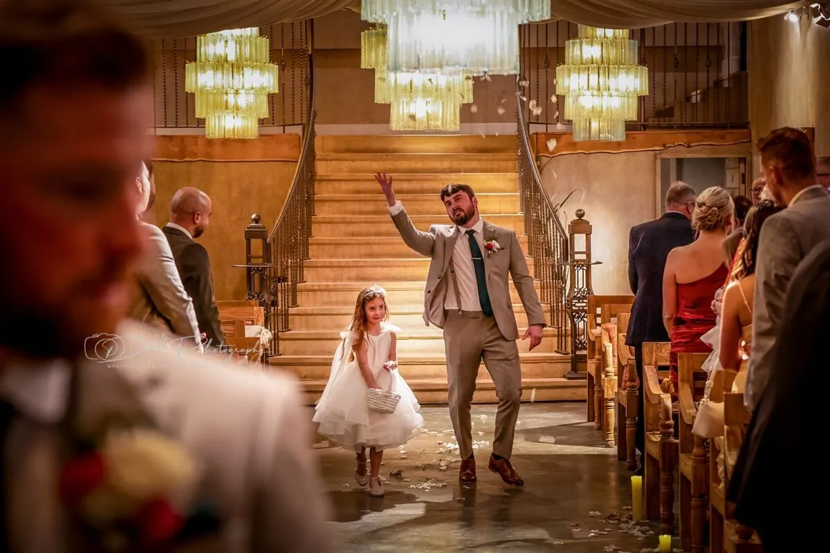 A groomsman playfully tosses confetti in the air while walking down the aisle with a flower girl carrying a small basket inside a venue with a grand staircase and chandeliers.