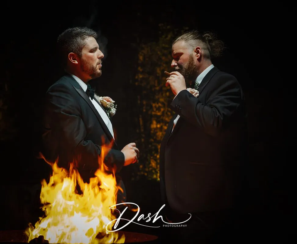 Two grooms in black tuxedos share a quiet evening moment together by a glowing outdoor fire pit while smoking cigars.