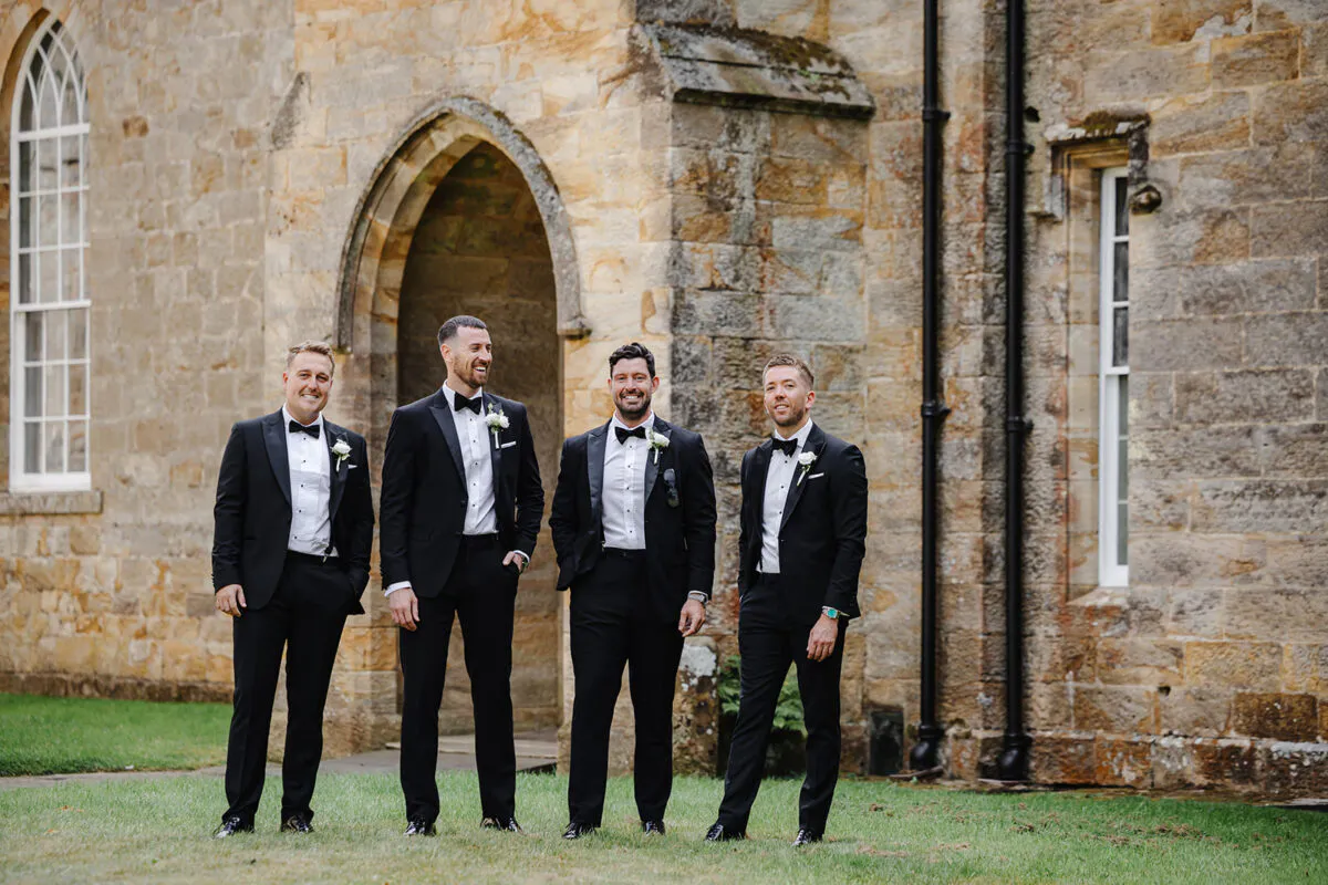 Four groomsmen wearing matching black tuxedos and bow ties stand smiling on the lawn outside a historic stone church.