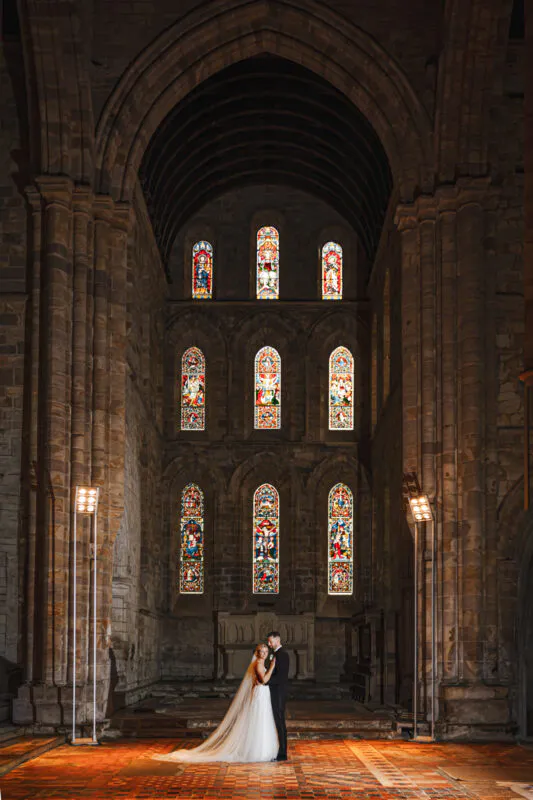 A newlywed couple stands small beneath the towering stone arches and magnificent stained glass windows of a grand cathedral.
