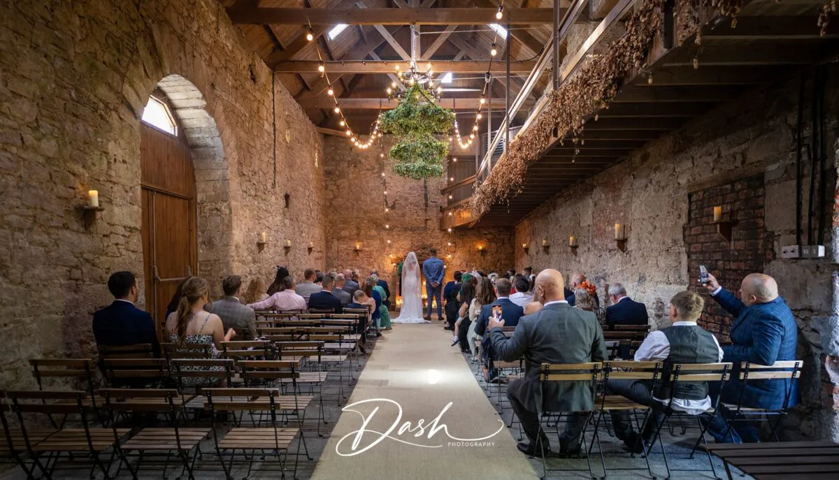 Guests seated on wooden chairs watch a wedding ceremony take place inside a rustic stone barn with exposed wooden rafters and hanging floral installations.