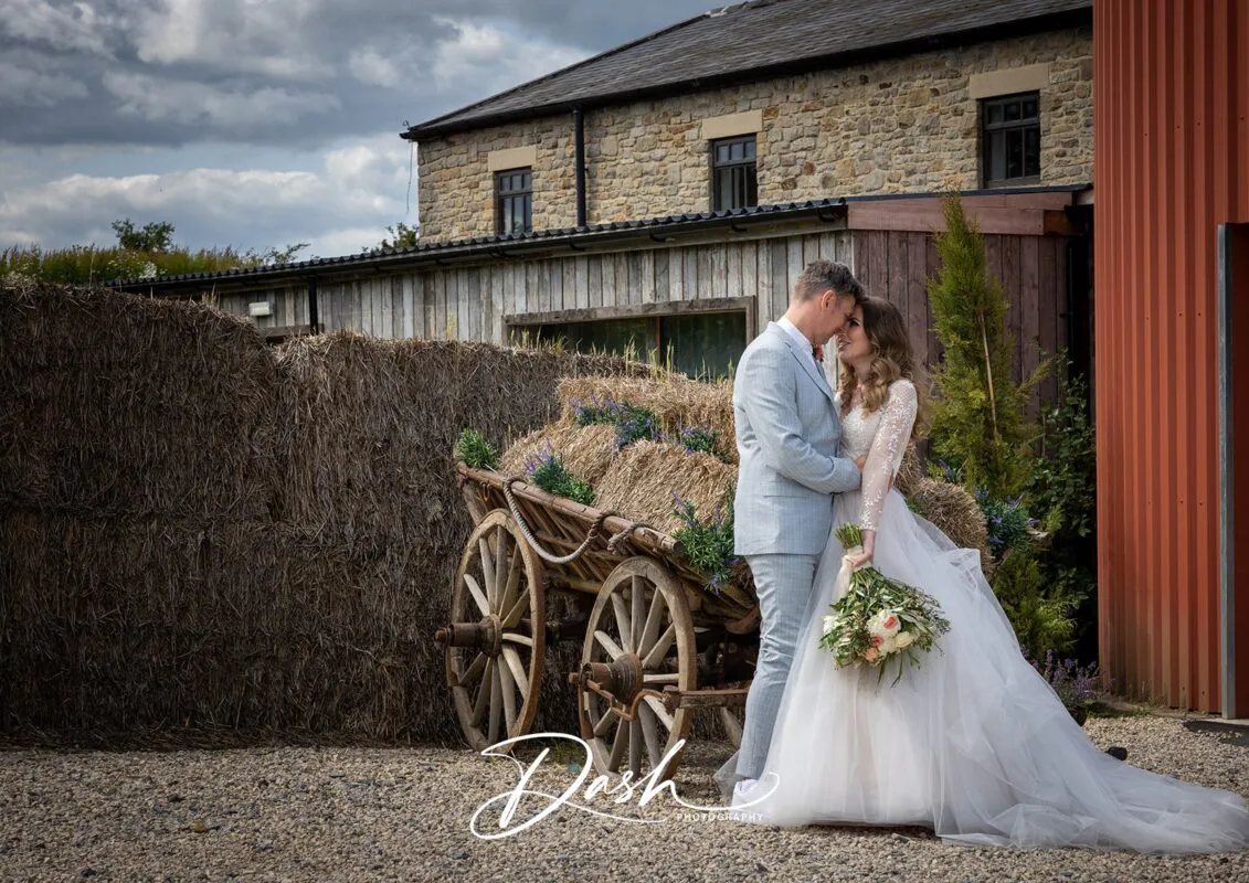 A newlywed couple embraces lovingly next to a rustic wooden cart filled with hay bales outside a traditional stone barn on their wedding day.