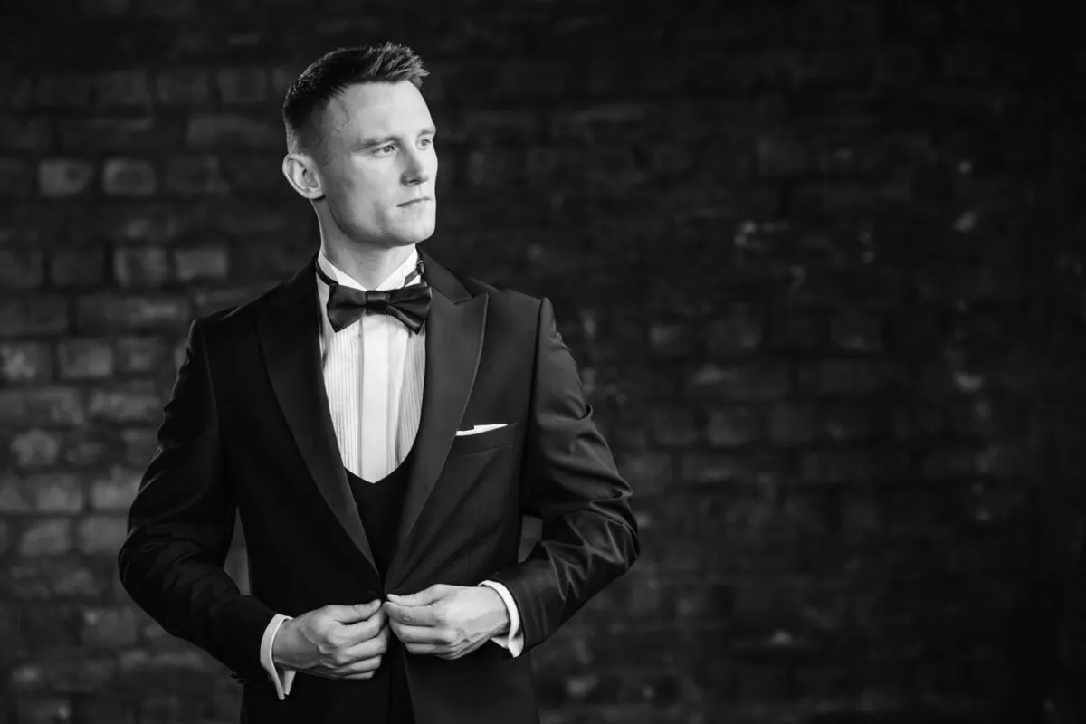 A stylish groom stands against an exposed brick wall, elegantly adjusting his black tuxedo jacket.