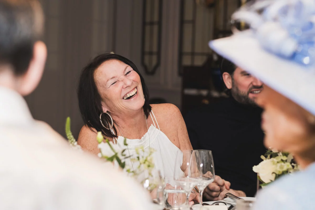 An older female wedding guest seated at a reception dinner table throws her head back in a joyful, open-mouthed belly laugh.