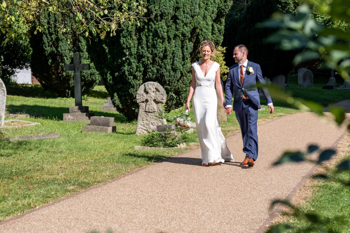 A newlywed couple walks hand-in-hand along a gravel path in a churchyard on a sunny summer day.