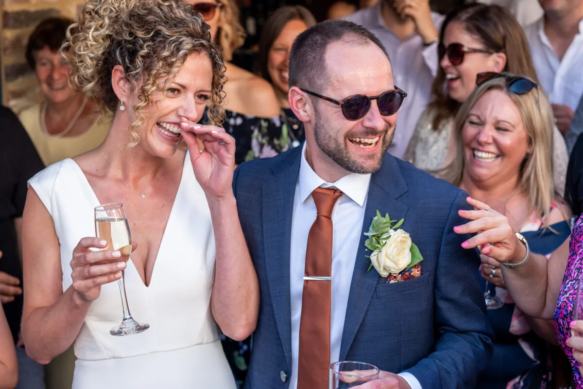 A laughing bride holding a glass of champagne stands next to her groom in a navy suit and sunglasses amidst a joyful crowd.