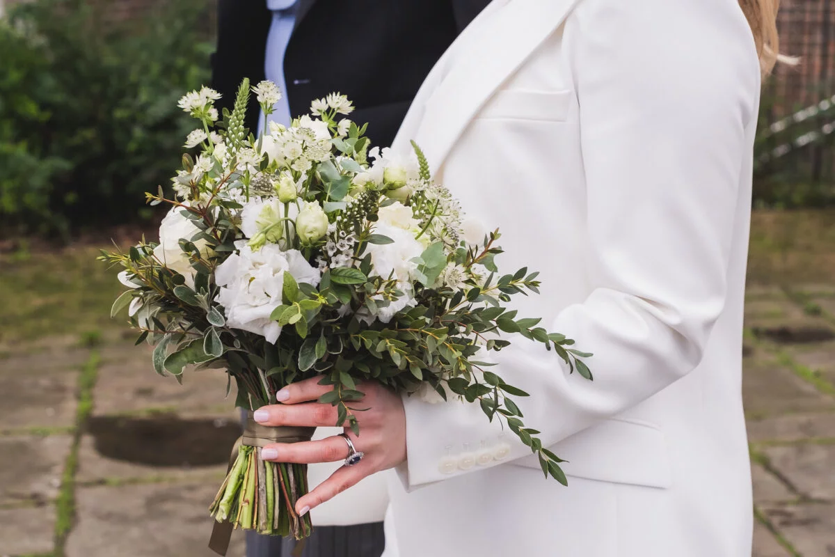 A bride wearing a white trouser suit holds a hand tied white and green bridal bouquet featuring delicate lisianthus and trailing eucalyptus.