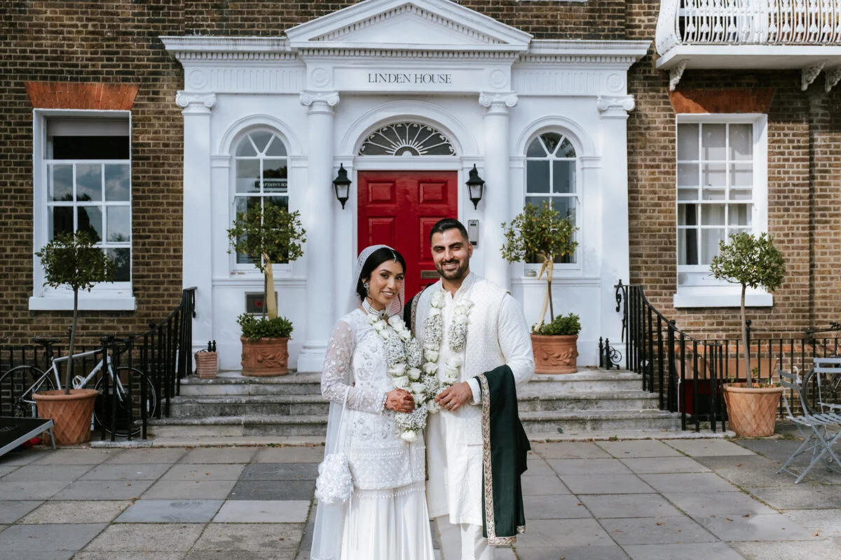 A newlywed couple poses together in front of the grand white portico and red door of Linden House, wearing traditional white wedding attire adorned with white floral garlands.