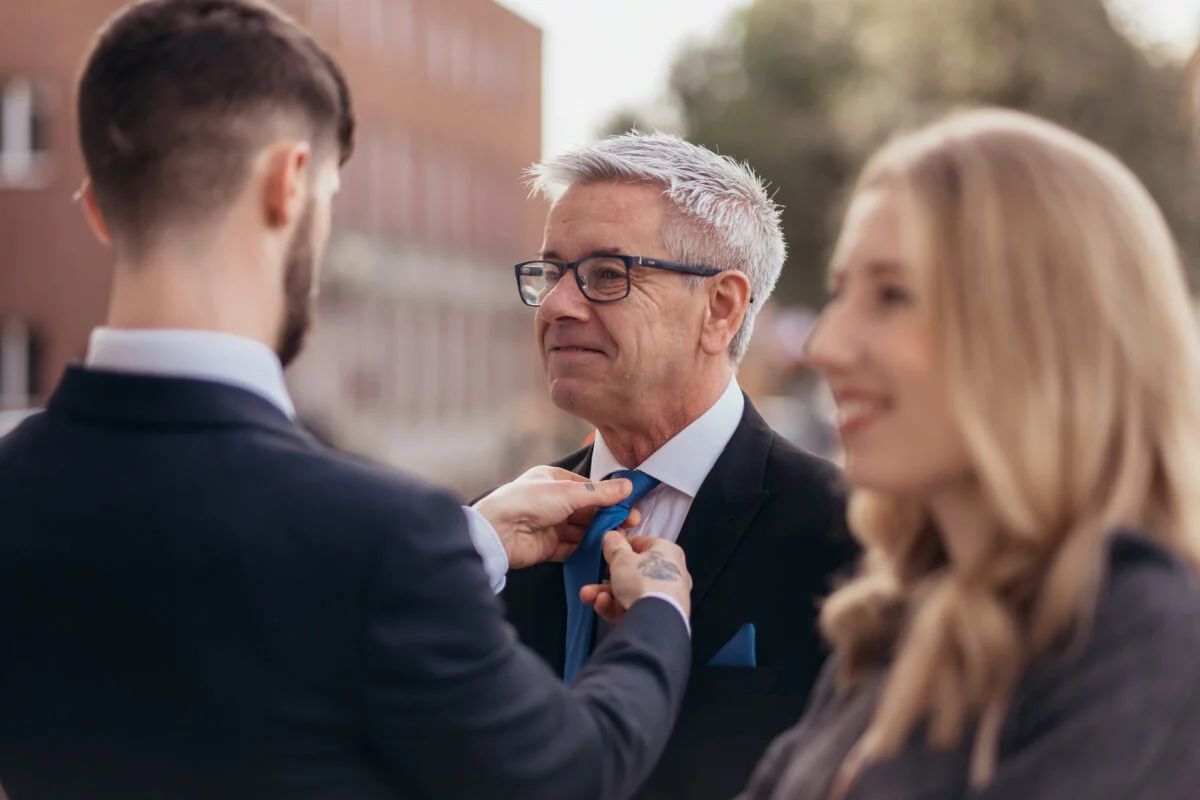 An older gentleman in a black suit has his blue tie adjusted by a family member outdoors, with a smiling blonde woman blurred in the foreground.