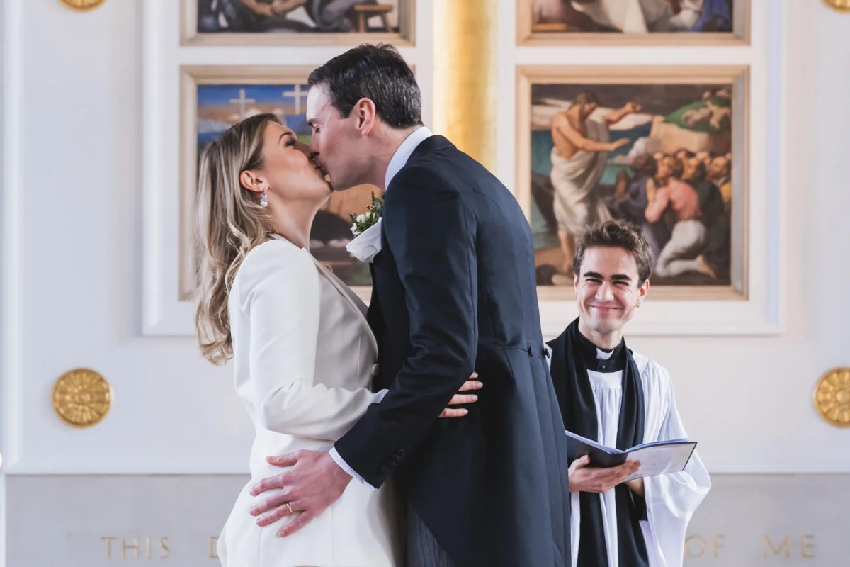 A newly married couple share their first kiss at the altar during a traditional church ceremony while a smiling official looks on.