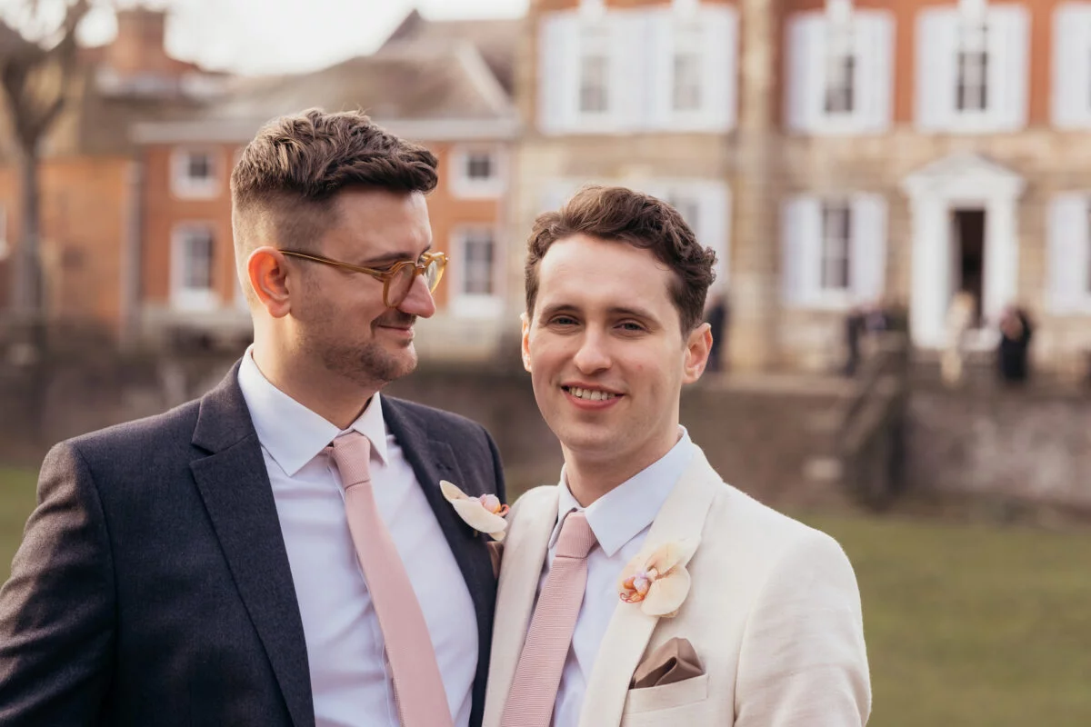 Two grooms in stylish suits stand together outside a historic manor house, sharing a tender look with matching pink ties and floral boutonnieres.