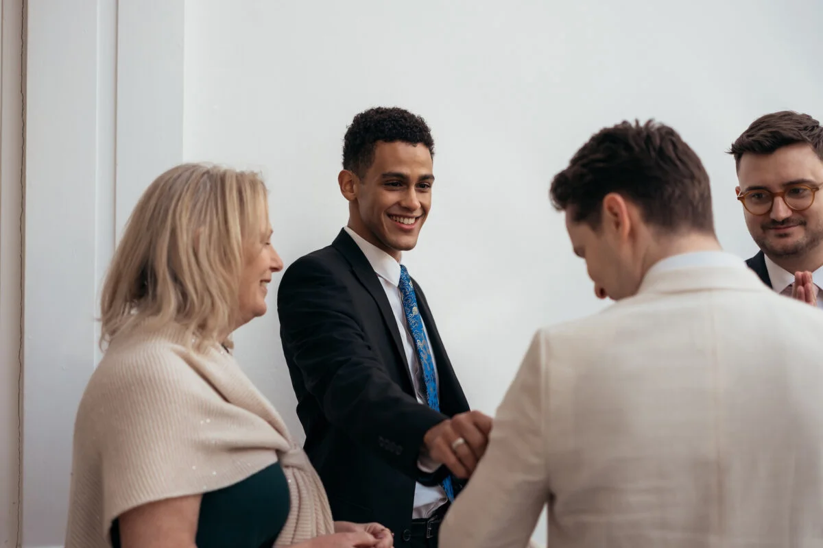 A smiling guest in a black suit shakes hands with a groom in a beige suit while wedding guests converse nearby.