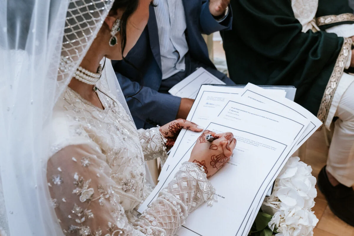 A south asian bride adorned with intricate henna on her hands signs the marriage certificate while wearing a beaded long-sleeved gown and pearl-embellished veil.